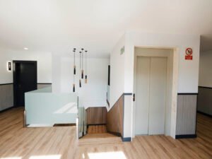 Rellano de la planta superior con puerta de ascensor, escaleras con barandilla de cristal y entrada a habitaciones en la Pensión Casa Cancela.