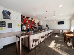 Barra de bar con encimera de granito, taburetes altos de madera y lámparas de estilo industrial. Se aprecia la máquina de café profesional y una pared decorada con temática de café.