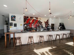 Barra de cafetería con encimera de mármol oscuro, taburetes de madera circulares y un original sistema de iluminación con cables rojos y bombillas colgantes sobre un fondo decorado.
