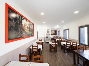 Salón de restaurante amplio y luminoso con mesas preparadas, sillas de madera rústica y grandes cuadros con marcos rojos que muestran paisajes rurales.