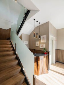Escaleras de madera con barandilla de cristal junto al mostrador de recepción rústico y ascensor.