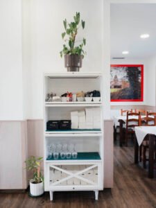 Mueble de servicio blanco en restaurante con copas de vino, servilletas y salsas, decorado con plantas naturales y vista al comedor con mesas preparadas al fondo.