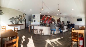 Vista panorámica del interior de una cafetería moderna con clientes en la barra, mesas de madera, iluminación de bombillas colgantes y un mural artístico de una planta de café en la pared.