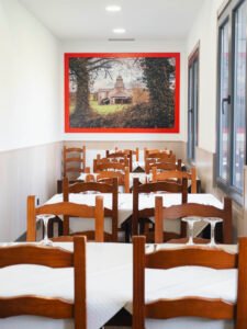 Comedor interior con hileras de mesas de madera preparadas con mantelería blanca y copas, presidido por un gran cuadro de un paisaje rural en la pared del fondo