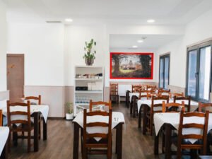 Comedor de restaurante luminoso con mesas de madera vestidas con manteles blancos, sillas rústicas, suelo de tarima y un cuadro decorativo al fondo.