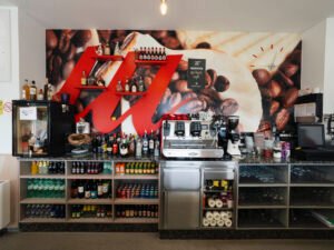 Zona de trabajo tras la barra de una cafetería con máquina de café espresso profesional, molinillos, estanterías con bebidas y un gran mural decorativo de granos de café al fondo