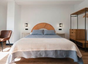 Interior de una habitación doble espaciosa en Casa Cancela (Palas de Rei), con un cabecero circular de mimbre, textiles en tonos azules y beige, y un armario abierto de estilo industrial.