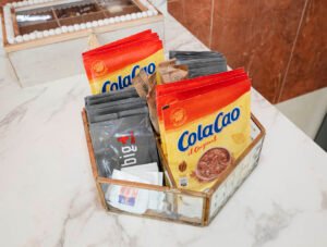 Detalle de bienvenida con sobres de cacao soluble, café descafeinado y azúcar para los huéspedes, presentados en una cesta sobre la encimera de la cocina del apartamento en Casa Cancela.